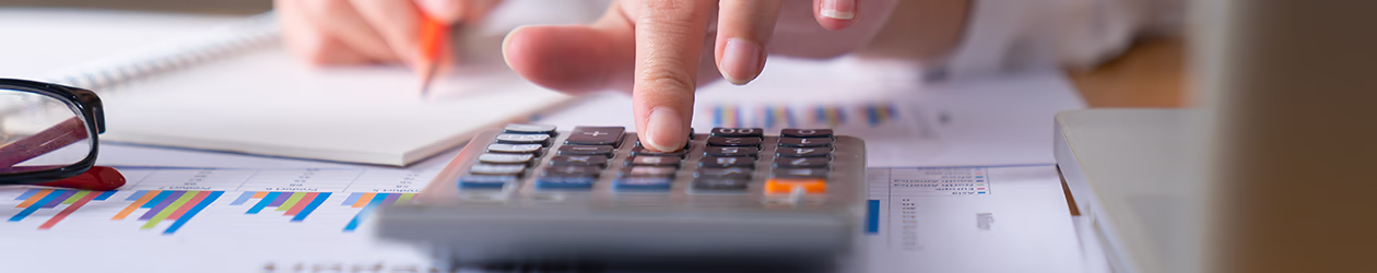 Person tippt auf einen Taschenrechner und notiert etwas mit einem Stift in einem Heft, umgeben von Unterlagen mit Diagrammen