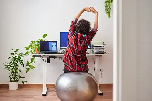 Eine Frau sitz auf einem Gymnastikball am Schreibtisch und streckt sich.
