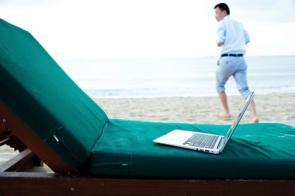 Ein Laptop liegt am Strand, im Hintergrund läuft ein Geschäftsmann.