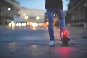 Ein Mann steht mit einem E-Scooter in der Dämmerung in der Stadt. Man sieht nur seine Beine und den E-Scooter von hinten.