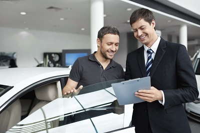 Autohändler und Kunde stehen gemeinsam bei einem Auto und betrachten lachend den Kaufvertrag.