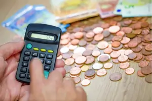 Nahaufnahme eines Taschenrechners, der von zwei Händen gehalten wird. Im Hintergrund liegt ein Haufen Kleingeld und einige Euroscheine