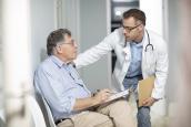Arzt hält eine Mappe und spricht mit einem älteren Patienten im Wartebereich.