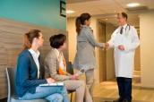 Arzt begrüßt eine Patientin, zwei weitere Patientinnen warten.