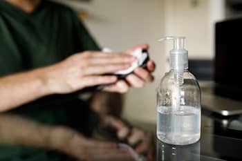 Eine Flasche mit Desinfektionsmittel steht auf einem Tisch, im Hintergrund reinigt eine Person ein Smartphone mit einem Tuch.