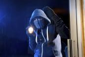 Einbrecher steht mit einer Taschenlampe an einer Wohnungstür und blickt hinein.