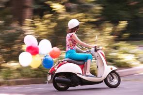 Eine Frau fährt auf einem Roller und zieht dabei Luftballons hinter sich her.
