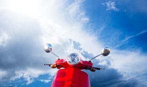 Ein rotes Moped steht vor blauem Himmel mit Wolken.