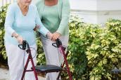 Seniorin geht mit einer Pflegeperson und mit Hilfe eines Rollators spazieren.