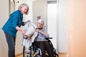 Angehörige hilft Seniorin im Rollstuhl in die Jacke.