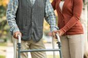 Senior geht mit Hilfe einer Pflegerin an einem Rollator.