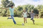 Mann, Frau und Kind spielen auf einer Wiese Fußball.