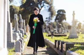 Eine junge Witwe mit Blumestrauß läuft über einen Friedhof.