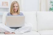 Eine Frau sitzt im Schneidersitz auf einer Couch und surft mit einem Laptop im Internet.