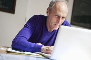 Einige Rentner zahlen ab April geringere Beiträge zur Pflegeversicherung. Älterer Mann sitzt am Laptop mit Papieren.