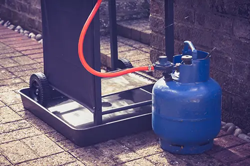 Eine blaue Gasflasche steht auf dem Boden vor einem Grill.