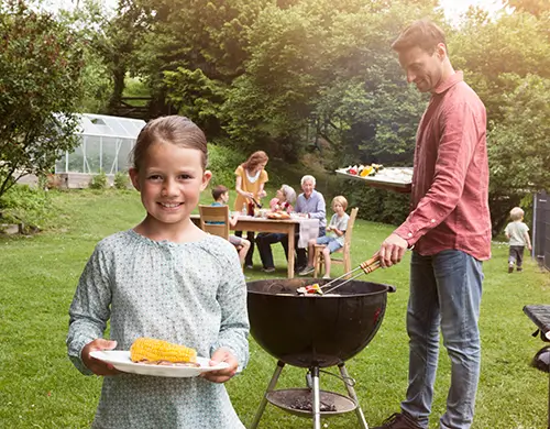 Eine Familie grill in ihrem Garten, im Vordergrund steht die junge Tochter mit einem Teller.