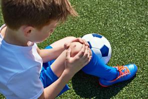 Junge mit aufgeschürftem Knie sitzt mit einem Fußball auf dem Rasen.
