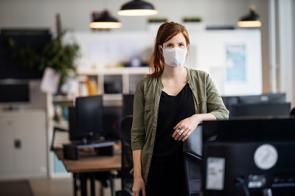 Junge Angestellte steht mit Maske im Großraumbüro