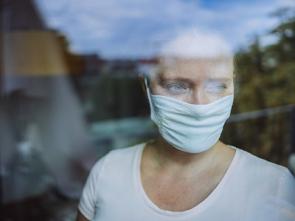 Frau schaut mit Maske während Corona-Pandemie aus dem Fenster.