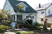 Ein Mann steht vor einem Haus, das von einem entwurzelten Baum schwer beschädigt wurde.