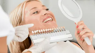 Eine Patientin beim Zahnarzt hält einen Handspiegel um ihre Zahnfarbe mit einem Modell zu vergleichen.