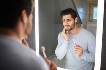 Ein Mann mit schmerzverzerrtem Gesicht steht beim Zähneputzen vor dem Spiegel.